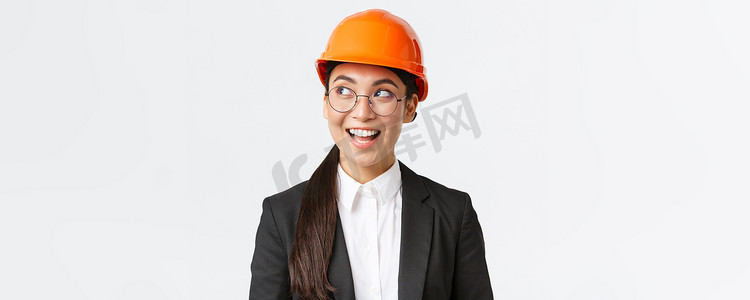 创意亚洲女建筑师的特写，心目中有有趣的想法，身穿西装和安全头盔的建筑工程师面带愉悦的微笑，站在白色背景下向左看