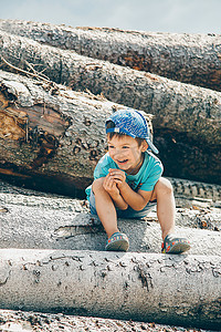 戴着棒球帽的学龄前男孩坐在木头上，开心地笑着