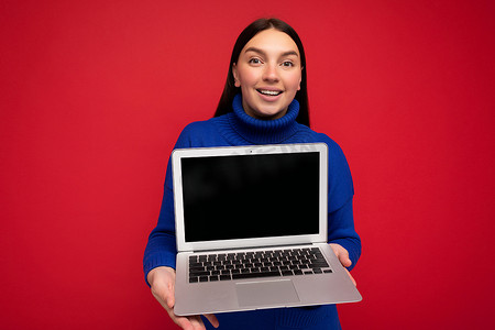 照片中，微笑迷人、迷人、快乐、美丽、黑发的年轻女子，直发，手持电脑笔记本电脑，看着相机，身穿蓝色毛衣，与红墙背景隔离