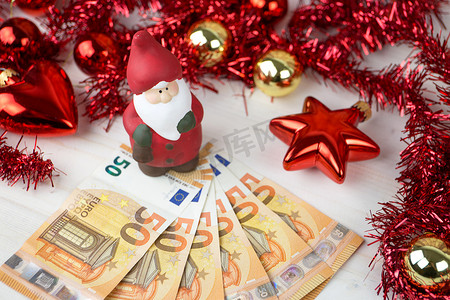 圣诞钱的商业理念：圣诞老人雕像在大约五十欧元纸币上，上面有红色和金色的小玩意，浅色木桌上有花环装饰