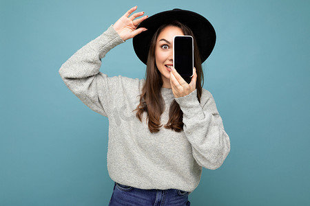 戴着黑帽和灰色毛衣的漂亮年轻微笑女人拿着手机，看着背景中突显的相机