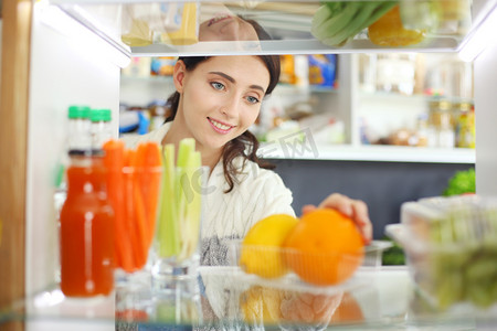 站在开着的冰箱旁边的女性画像，冰箱里装满了健康食品、蔬菜和水果。