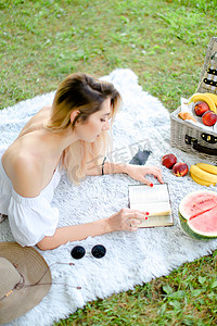年轻的女孩在看书，躺在格子上，靠近水果和帽子，背景是草。