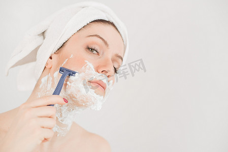 头戴毛巾、脸上剃须泡沫的快乐白人女性拿着白色背景上的剃刀