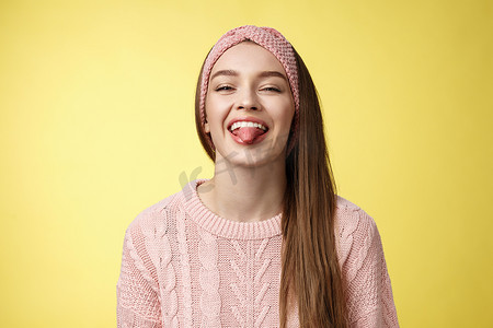 积极有趣的​​可爱风趣迷人的年轻欧洲女孩穿着毛衣，针织时髦头带笑着开玩笑地吐舌头，嘲笑朋友在黄墙上享受阳光明媚的日子