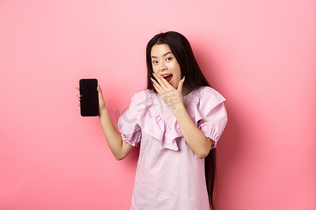 开朗的亚洲少女展示空的手机屏幕，笑着用手捂住嘴，站在粉色背景下的裙子