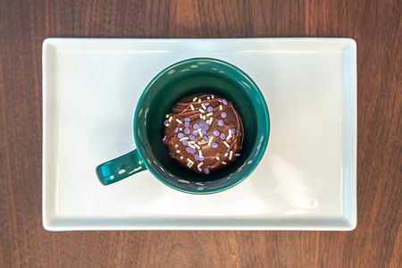 关闭热可可炸弹的顶视图，上面覆盖着毛毛雨的棕色巧克力和紫色和白色的洒水装置，放在木桌上的矩形服务盘上的绿色咖啡杯或杯子内。