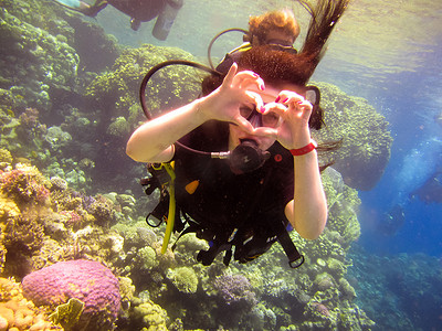 珊瑚可爱摄影照片_可爱的女潜水员用手势表达爱意，与合作教练一起在手指上表达出一颗心，在埃及沙姆沙伊赫的红海珊瑚礁中安全游泳