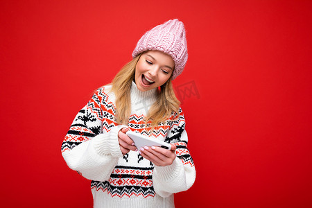 照片中，美丽微笑的年轻金发女子戴着温暖的针织帽和冬季温暖的毛衣，站在红色背景中，通过智能手机看着设备显示屏，与世隔绝地玩游戏