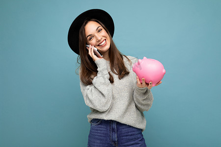 快乐积极微笑的年轻迷人黑发女性的肖像，身穿休闲灰色毛衣，头戴黑色帽子，蓝色背景上孤立，空间空旷，手持粉红猪便士银行，在网上用手机说话