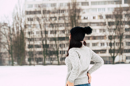 冬季穿着灰色针织毛衣和帽子站在街上的年轻女子的后视图。