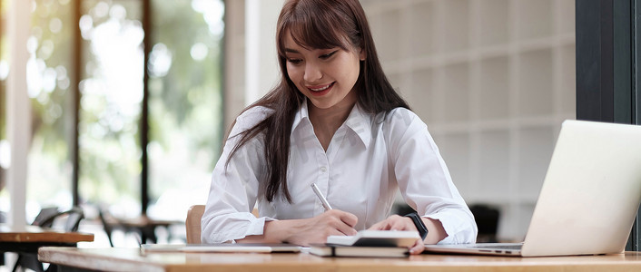 一个穿着白衬衫的年轻开朗的女孩微笑着，微笑着写下笔记，为学生在笔记本电脑桌面桌上进行高管培训
