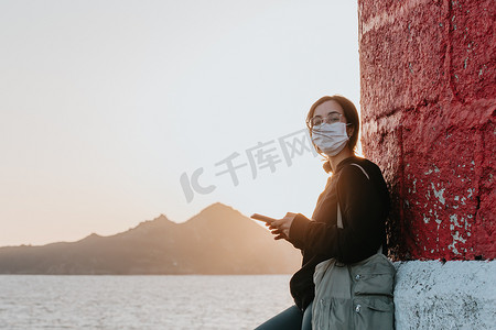 年轻女子在西班牙海岸用他的手机，以巨大的日落为背景，同时使用面具放松概念