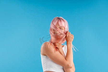 身穿白衬衫的微笑女模特玩着光滑的粉红色直发，在蓝色工作室背景下摆出孤立的姿势时遮住脸