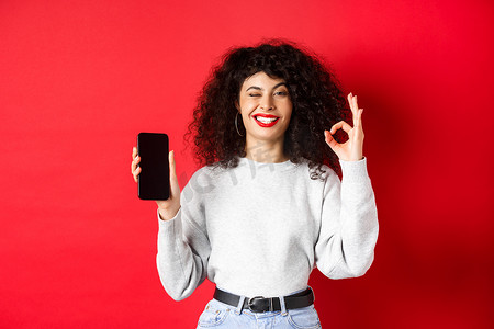 有智能手机的迷人女性，显示好的标志和空的电话屏幕，推荐购物应用程序，站在红色背景下