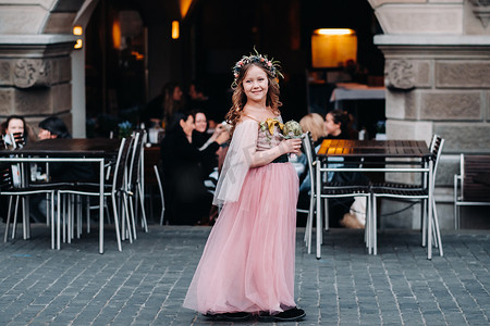 粉色裙摄影照片_一个穿着粉红色公主裙的小女孩手里拿着一束花穿过苏黎世老城。瑞士城市街道上一个穿着粉红色连衣裙的女孩的肖像