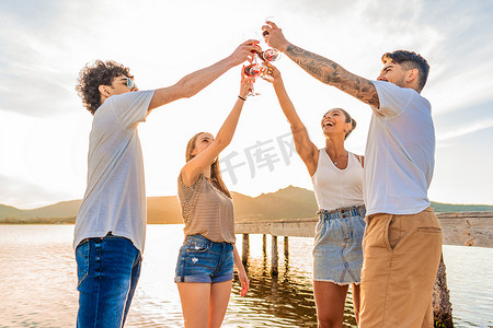 一群多种族的快乐朋友在日落时分在海滩上用红酒举杯，在背光的阳光下碰杯。