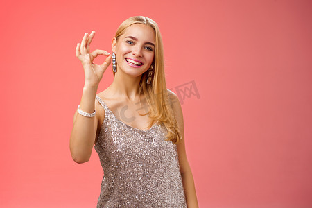 乐观迷人、时尚的金发女性穿着银色连衣裙，表现得很好，手势微笑，广泛给予认可，比如推荐酷店购买服装生日派对，站在红色背景上