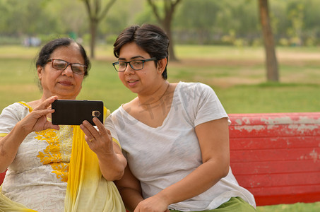 印度新德里摄影照片_印度新德里一个公园的红色长凳上，年轻的印度女孩和她的印度老母亲看着手机，忙着自拍