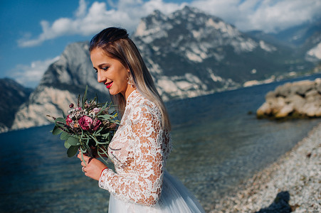 一个穿着漂亮的白色连衣裙的女孩沿着加尔达湖的堤岸散步。一个女人在意大利的一座山和湖的背景下被拍照。Torbole