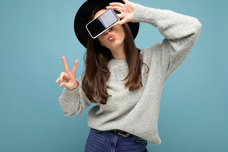 戴着黑帽和灰色毛衣的漂亮年轻女人拿着手机，手机上展示着背景中突显亲吻嘴唇的智能手机