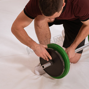重量训练摄影照片_给杠铃年轻人增加重量，跪在膝盖上，换黑色和绿色的盘子，重量训练概念的设备。