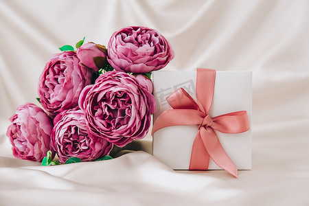 白色礼物盒，白色丝绸面料背景上有粉色丝带和紫罗兰色牡丹花。