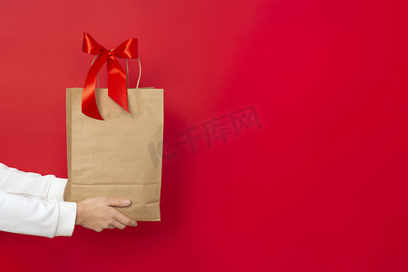 棕色海报摄影照片_女性手持大礼品袋，由棕色牛皮纸制成，红色背景上有红色蝴蝶结