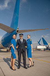 欢快的飞行员和两位漂亮的空姐站在飞机前，对着镜头微笑的全长镜头