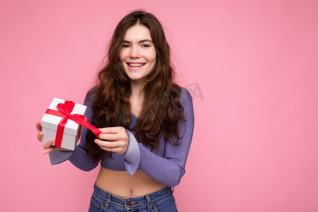 拆圣诞节礼物摄影照片_照片中，迷人的正面微笑的黑发卷发年轻女性被隔离在粉红色背景墙上，身穿紫色衬衫，手拿带红丝带的白色礼盒和拆箱礼物，看着相机