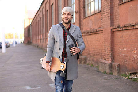英俊的年轻人穿着灰色外套和帽子走在街上，使用长板。