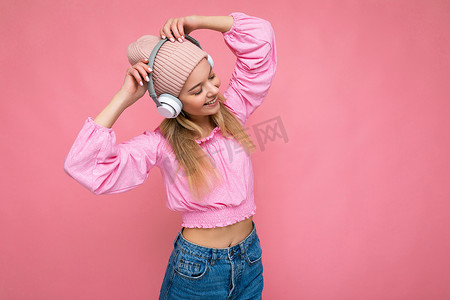 美丽快乐的微笑年轻金发女子穿着粉红色衬衫和粉红色帽子，隔离在粉红色背景墙上，戴着白色无线蓝牙耳机，听酷音乐，玩得开心，跳舞