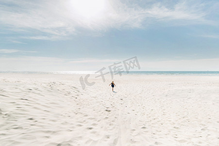 雪白的摄影照片_一个孩子在阳光明媚的天气里沿着白色的沙滩跑向波罗的海。一个小女孩跑过雪白的沙滩迎接大海。立陶宛