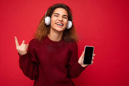 一张迷人的正面微笑的深色卷发年轻女子身穿深红色毛衣，与红色背景墙上隔开，戴着白色无线耳机，听音乐，展示手机，屏幕空空如也，可以看到相机和节目