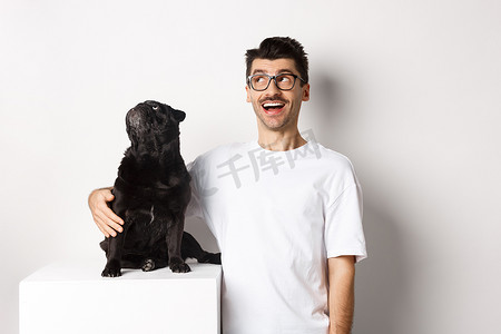 戴眼镜的年轻人惊讶地拥抱着他的狗、宠物主人和哈巴狗，盯着左上角的促销优惠，站在白色背景上