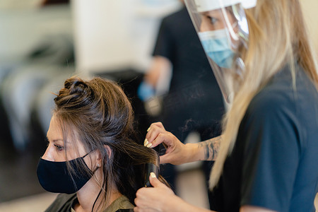 烫发插画摄影照片_美发师戴着面具，在沙龙里用烫发器梳理客户的头发。