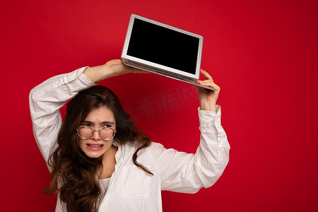 笔记本电脑俯视摄影照片_照片拍摄的滑稽美丽愤怒的黑发年轻女子拿着电脑笔记本电脑与空监控屏幕与模拟和复制空间穿着白衬衫俯视着与红墙背景隔离的一侧