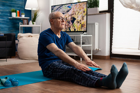 老人坐在客厅的瑜伽垫上伸展腿部肌肉
