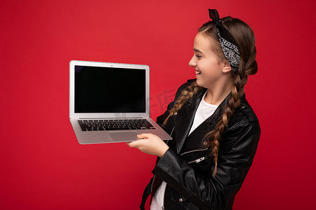 侧面照片：美丽快乐的微笑女孩留着深色辫子拿着电脑笔记本电脑穿着黑色夹克和头巾隔离在红墙背景上看着上网本显示器