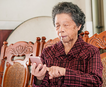 亚洲老年妇女坐在现代智能手机上浏览某些东西，与家里的其他人建立联系，生活技术，特写