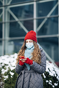 戴着面具的快乐的白人美丽卷发女性站在街上浏览智能手机。