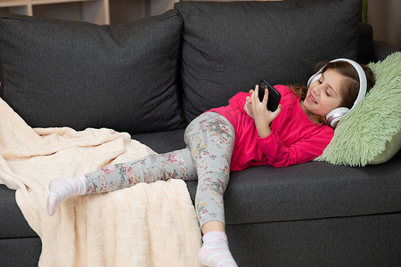 戴着无线耳机的快乐小女孩在沙发上大笑，使用移动在线播放器应用拿着手机听着最喜欢的音乐，在家里戴着耳机享受宁静的心情
