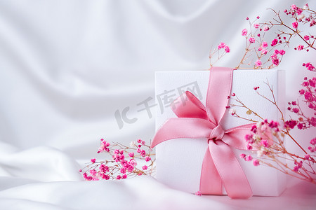 小花装饰摄影照片_白色礼物盒，带粉红丝带和白色丝绸织物背景上的粉红色小花。