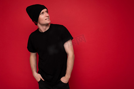 帅气积极的年轻人站在红色背景墙上，身穿黑色 T 恤，戴着黑色帽子，看着旁边