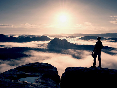 徒步旅行者的剪影站在悬崖上，看着迷雾笼罩的山谷到太阳。
