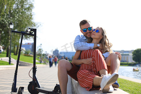 开心驾驶摄影照片_可爱的情侣在度假时玩得开心驾驶电动滑板车，从驾驶中休息，坐在河岸上