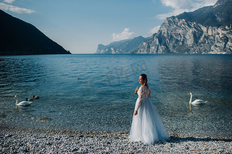 一个穿着漂亮的白色连衣裙的女孩沿着加尔达湖的堤岸散步。一个女人在意大利的一座山和湖的背景下被拍照。Torbole
