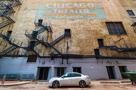 橙色的汽车摄影照片_芝加哥 — 2017 年 3 月，美国伊利诺伊州：芝加哥剧院墙的广角视图，有消防楼梯、门、蓝色背景上带有橙色箭头的手写涂鸦标志、停在街上的汽车
