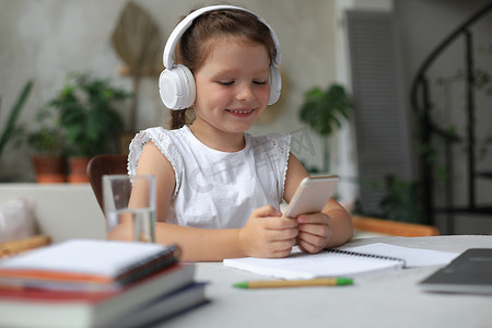 初等教育摄影照片_戴耳机的聪明小女孩在智能手机上在线上课，戴耳机的小孩在互联网上学习，在隔离期间举行网络会议或上课。