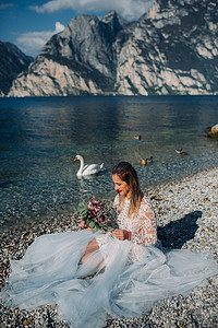 加衣服摄影照片_一个穿着漂亮的白色裙子的女孩坐在加尔达湖的堤岸上。一个女人在意大利的山湖背景下被拍到。Torbole